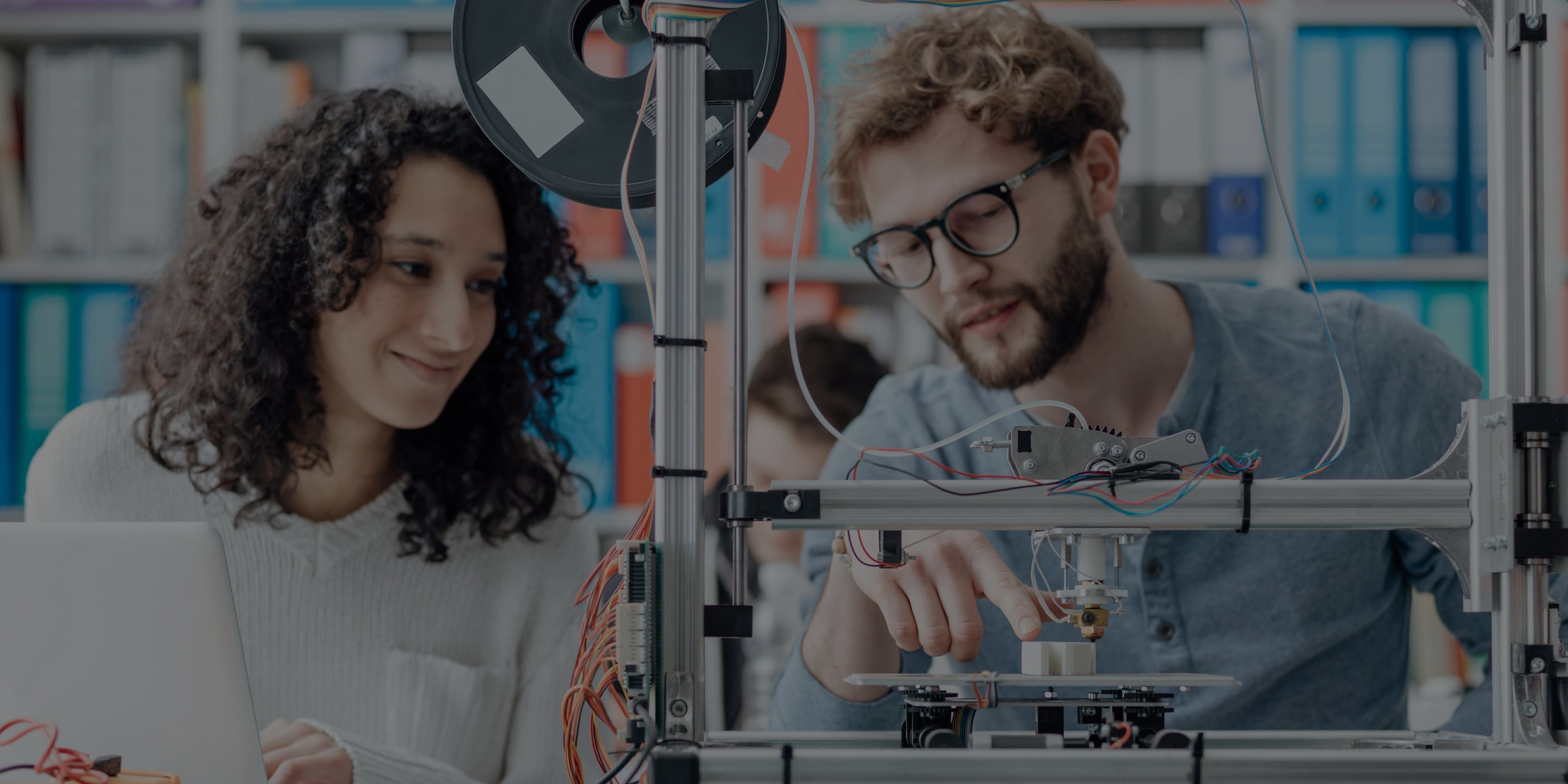 Two people working together on a 3D printer with electronic components and laptop in a lab setting.