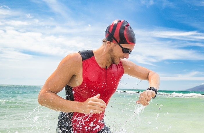 スポーツウォッチを着けて、水しぶきを上げながら海から駆け出している赤いトライアスロン･スーツを着たアスレチック･スイマー。