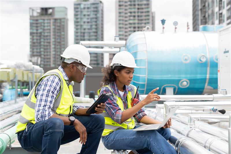 赤いヘルメットと黄色の安全ベストを着用した2人の作業員が、工場内で大型産業用ロボットを操作および点検しています。