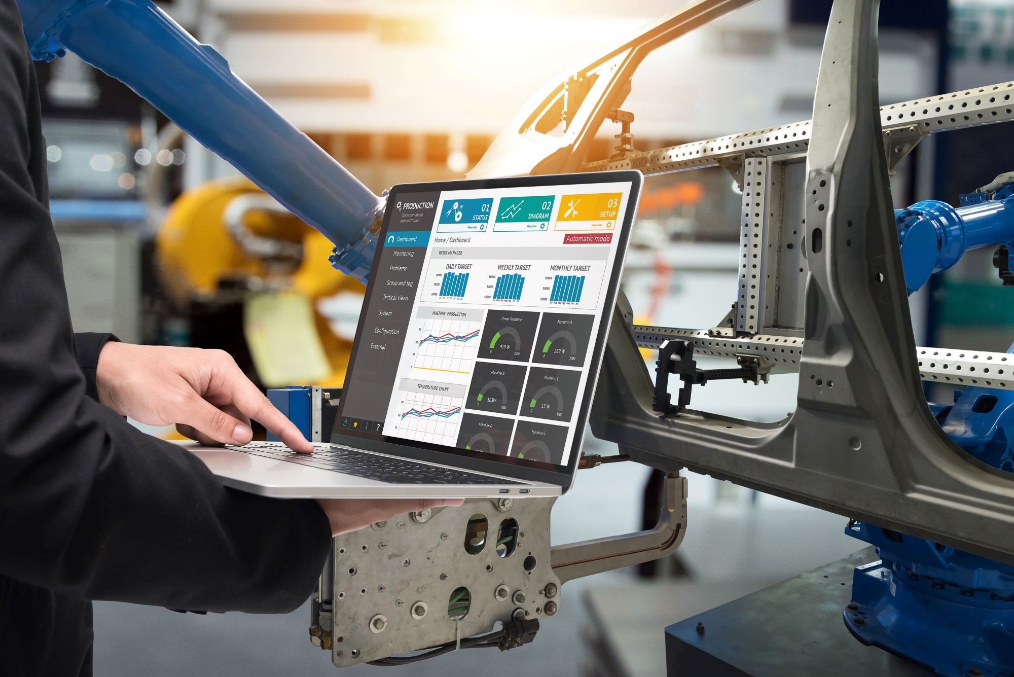A person holding a laptop displaying data analytics and charts, standing next to a robotic arm working on a car frame in an industrial setting. 