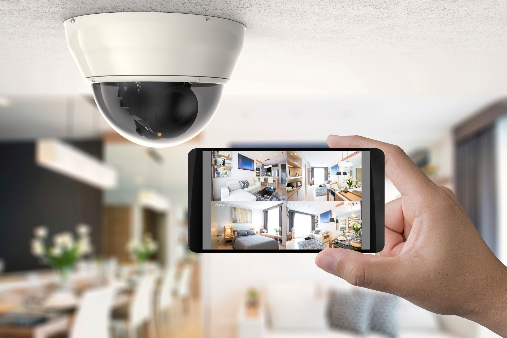 A hand holding a smartphone displaying a multi-camera home surveillance feed, with a white dome security camera mounted on the ceiling in the background. 