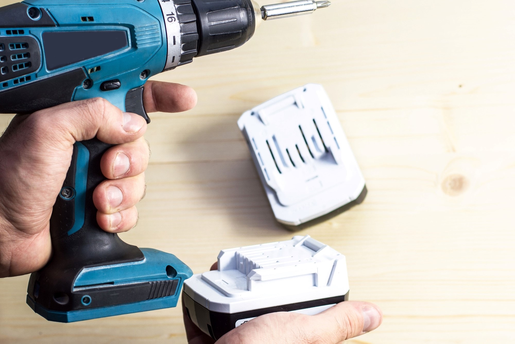A person’s hand holding a blue cordless power drill, with the other hand holding a white battery pack, preparing to insert or remove the battery on a light wooden surface. 