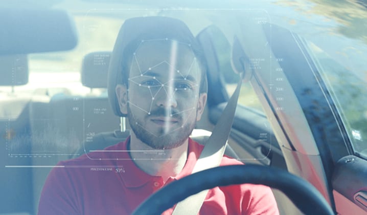 A person wearing a red shirt is seated in the driver's seat of a car. The image suggests the use of face authentication technology. 