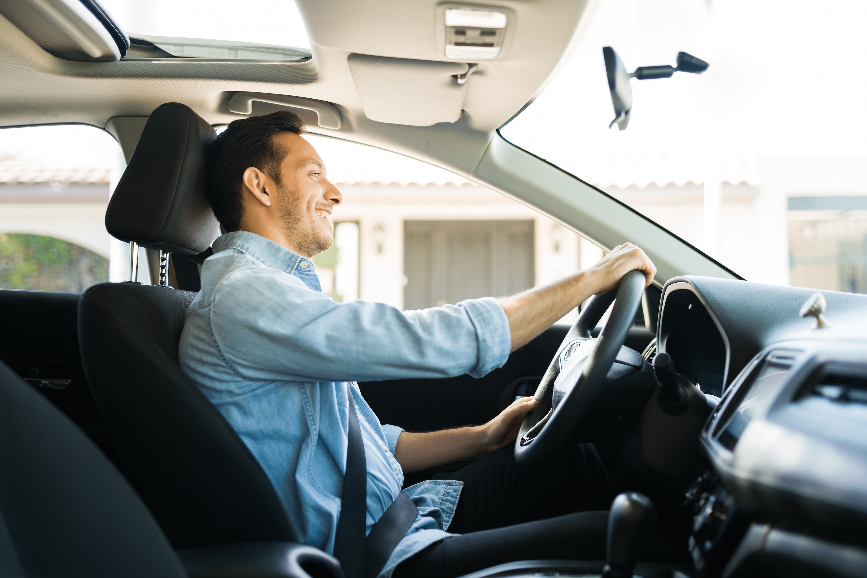 A person in a blue suit sits in the driver's seat of a car, holding the steering wheel, with sunlight streaming through the windows. 