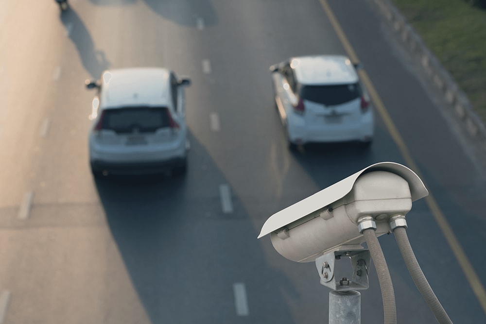 Image of a surveillance camera mounted above a road, monitoring traffic with two cars visible on the highway below.