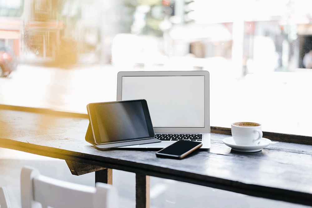 Laptop, tablet, and smartphone on a table next to a cup of coffee in a café.