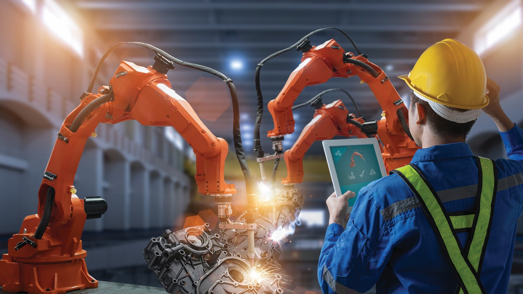 Engineer monitoring industrial robotic arms with a tablet in a factory. 