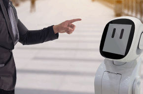 Person pointing at a small white humanoid robot with a digital face display.