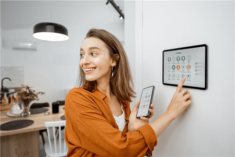 Woman using a wall-mounted smart home control panel while holding a smartphone in a modern kitchen.