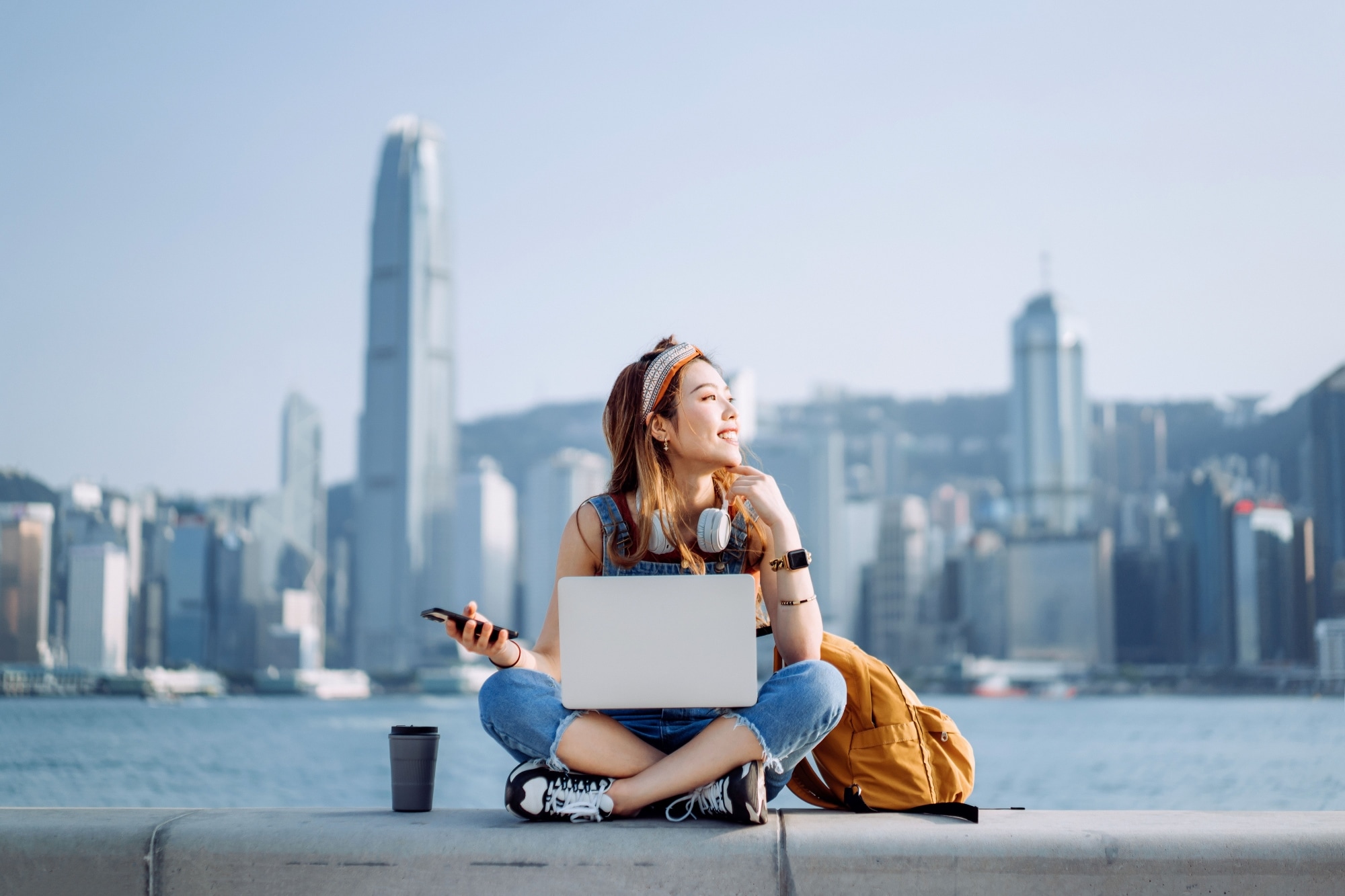 A person sitting cross-legged outdoors with a laptop on their lap, holding a smartphone, wearing headphones around their neck, with a city skyline and tall buildings in the background. 
