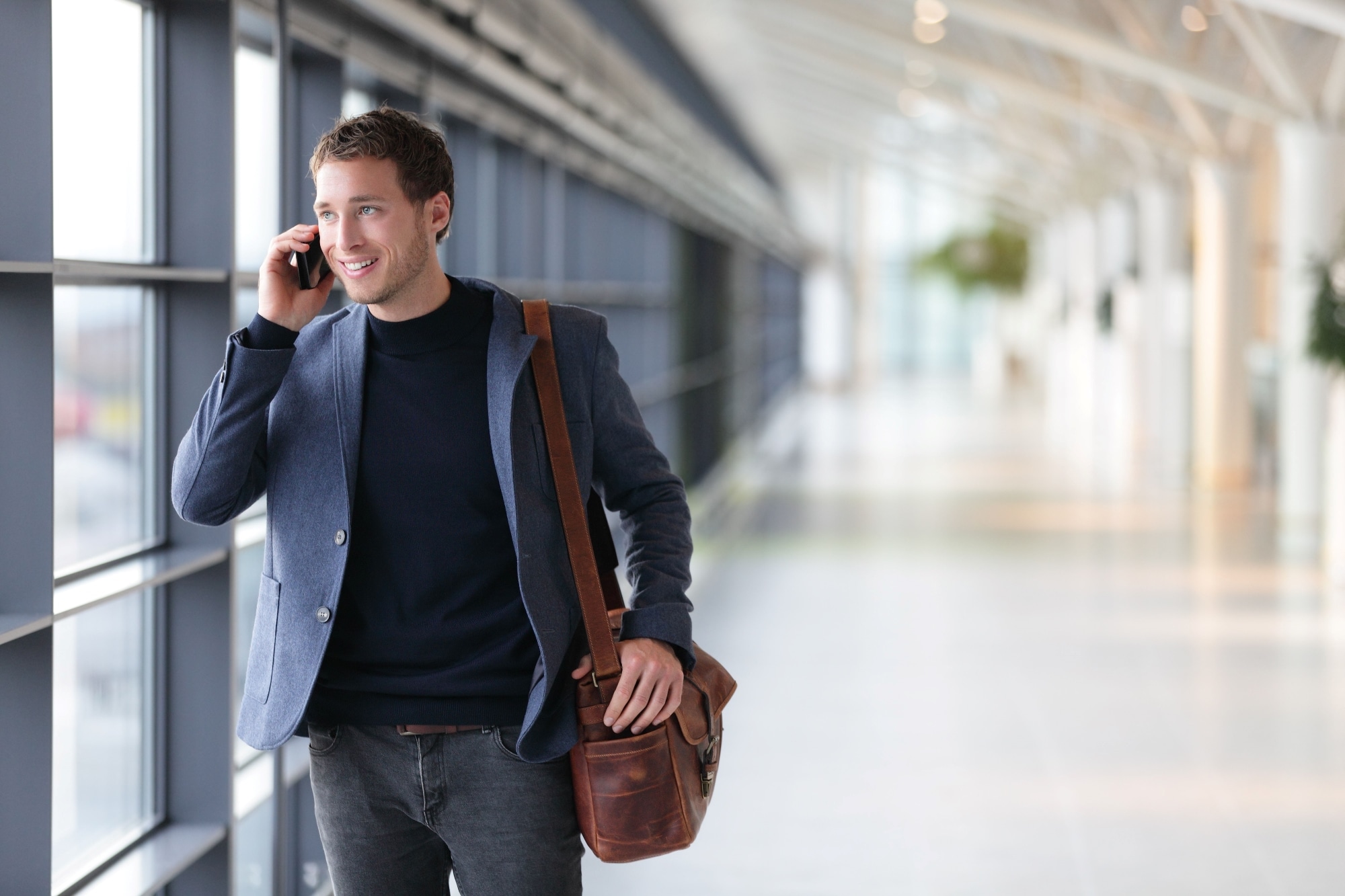 A person walking in a bright, modern corridor while talking on a smartphone, carrying a brown leather shoulder bag.