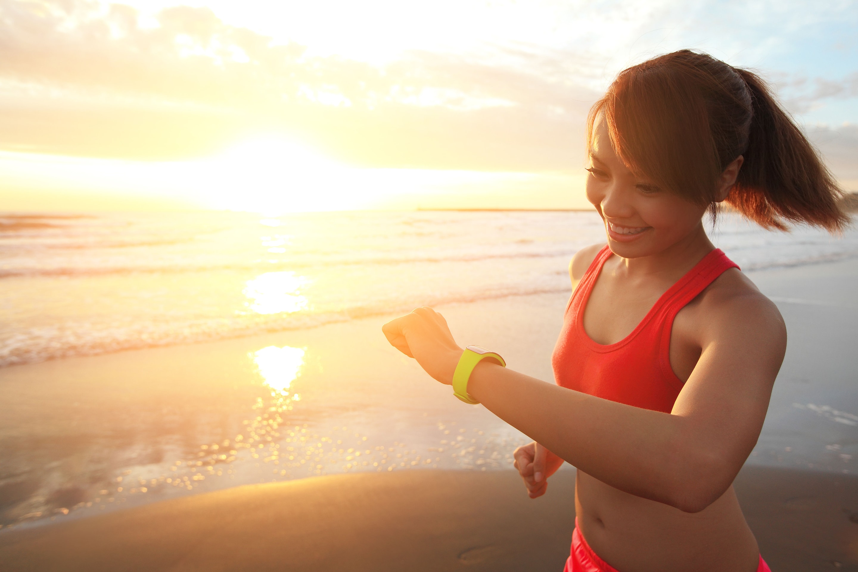 A person in a red sleeveless top is running on the beach at sunset, wearing a connected watch.