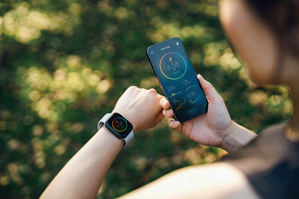 A person outdoors holding a smartphone displaying a fitness app while wearing a smartwatch on their wrist.