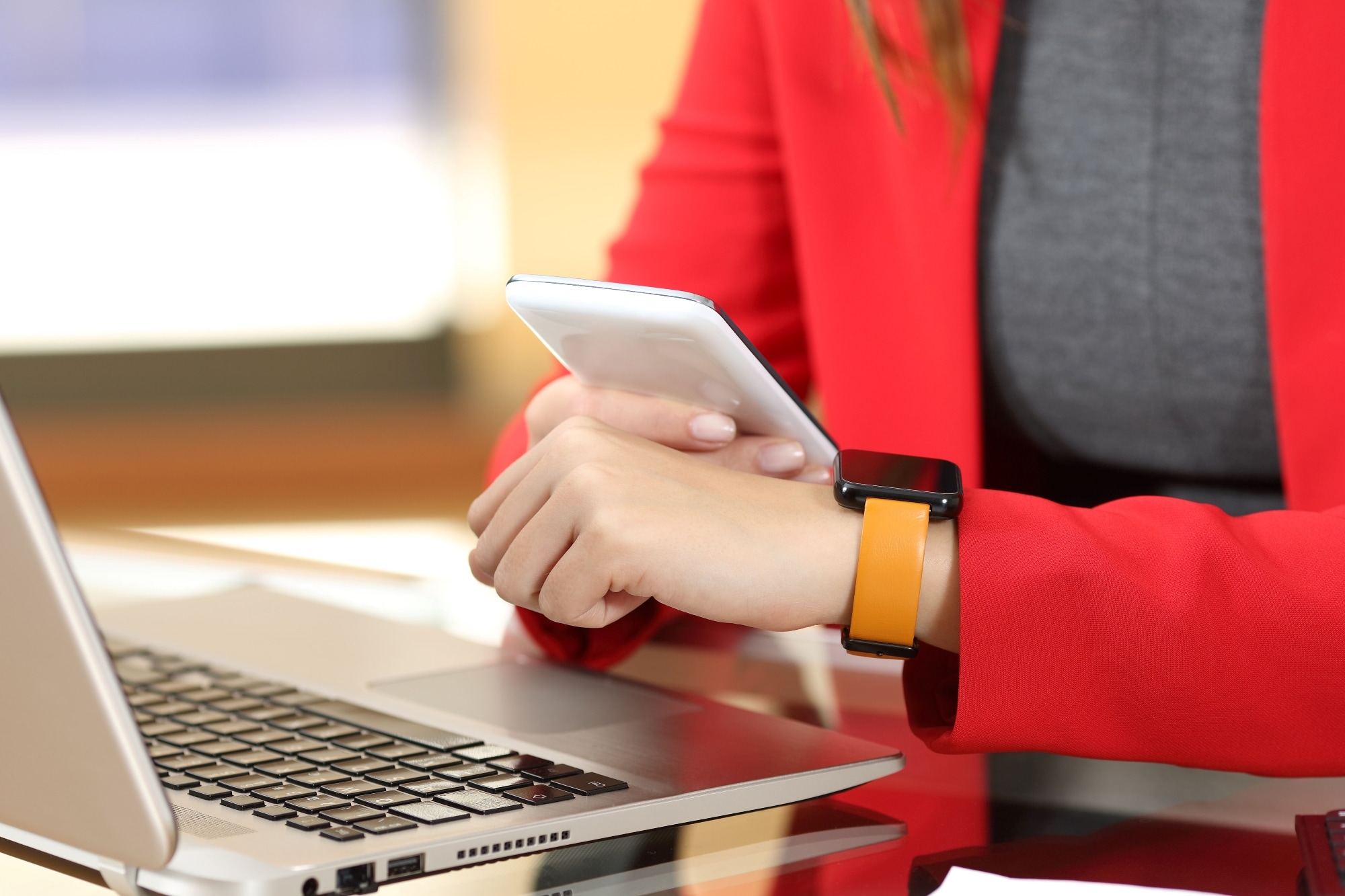 A person wearing a red jacket and an orange smartwatch, holding a smartphone near an open laptop.