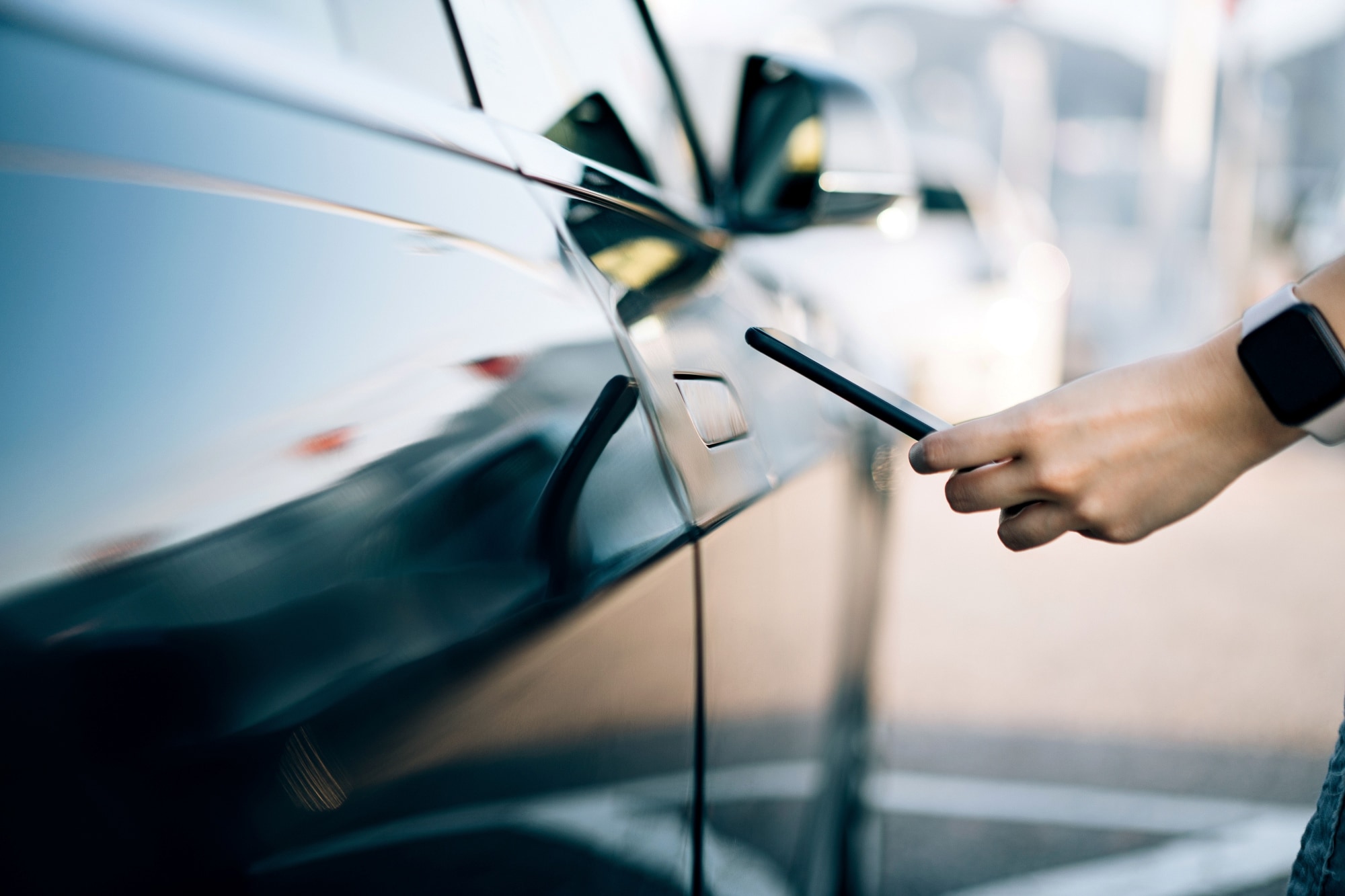 A person holding a smartphone near a black car door, using the phone to unlock the vehicle. 
