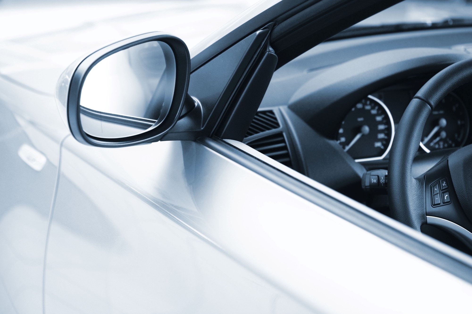 A close-up view of a silver car’s side mirror and part of the interior dashboard, including the steering wheel and instrument panel. 